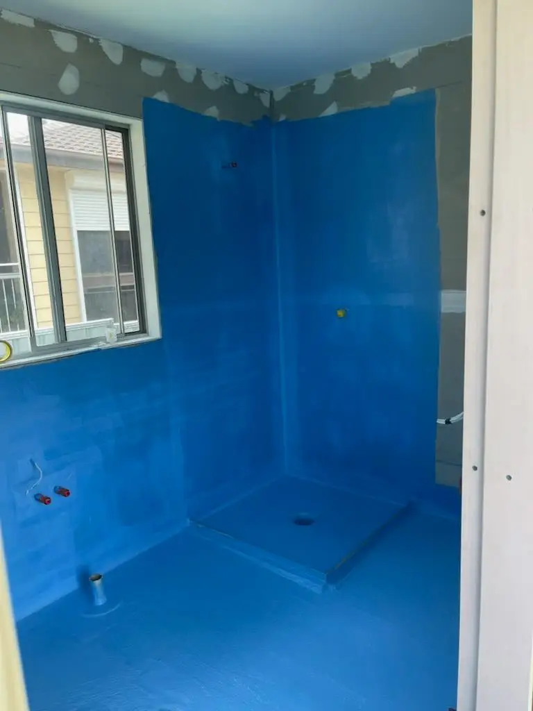 Blue waterproofing membrane applied to a bathroom floor and shower walls with the shower base falls formed and the hob turned up